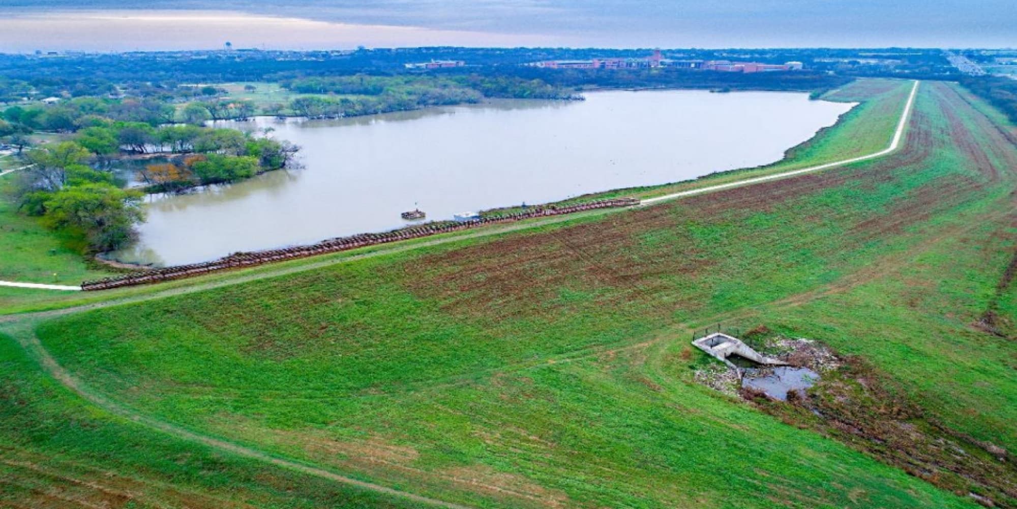 Aerial view of a grass-covered flood control embankment holding back its reservoir, with a Texas city skyline on the horizon.