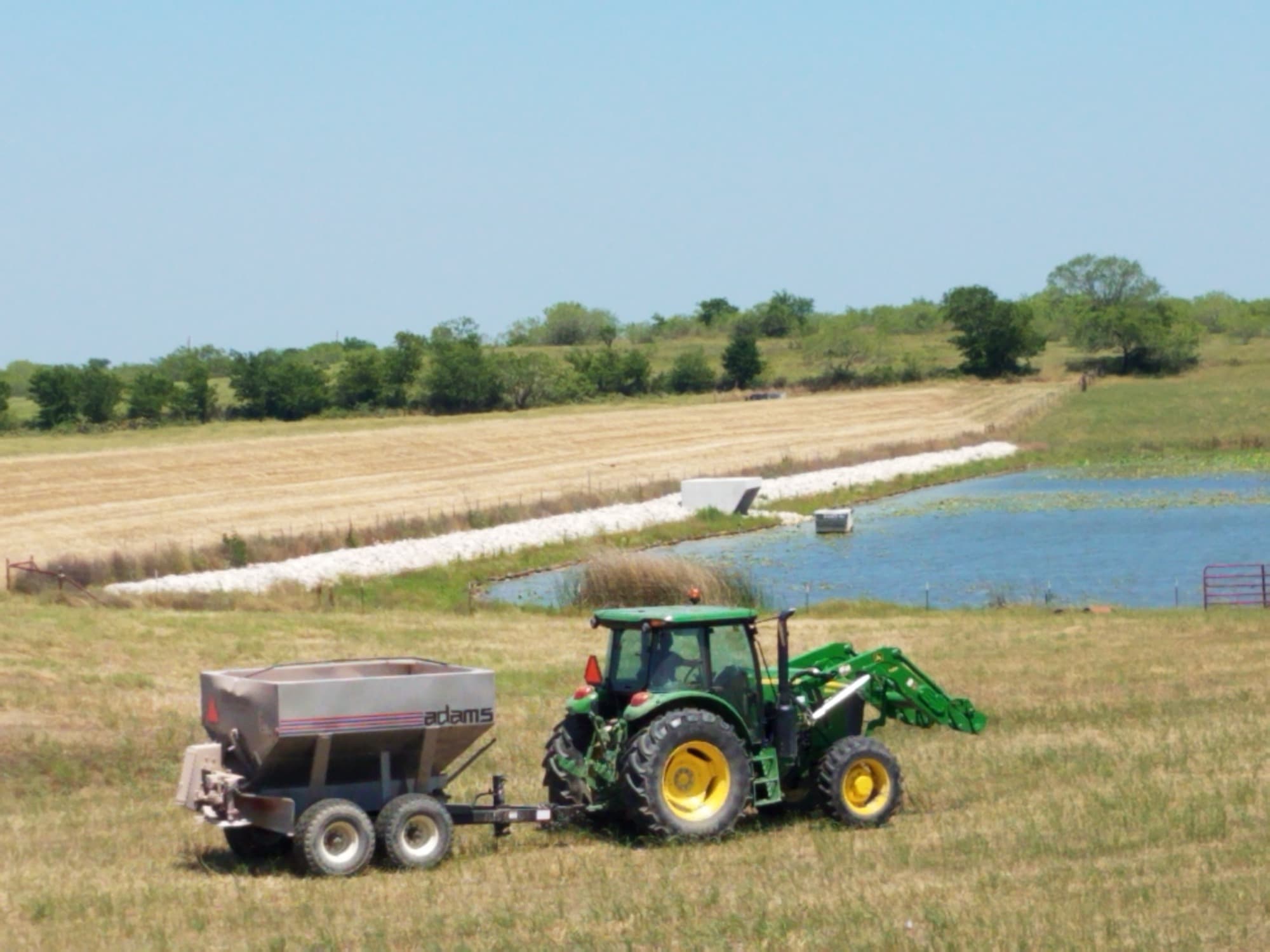 A tractor and spreader working the grassed slope of a flood control embankment beside its reservoir.