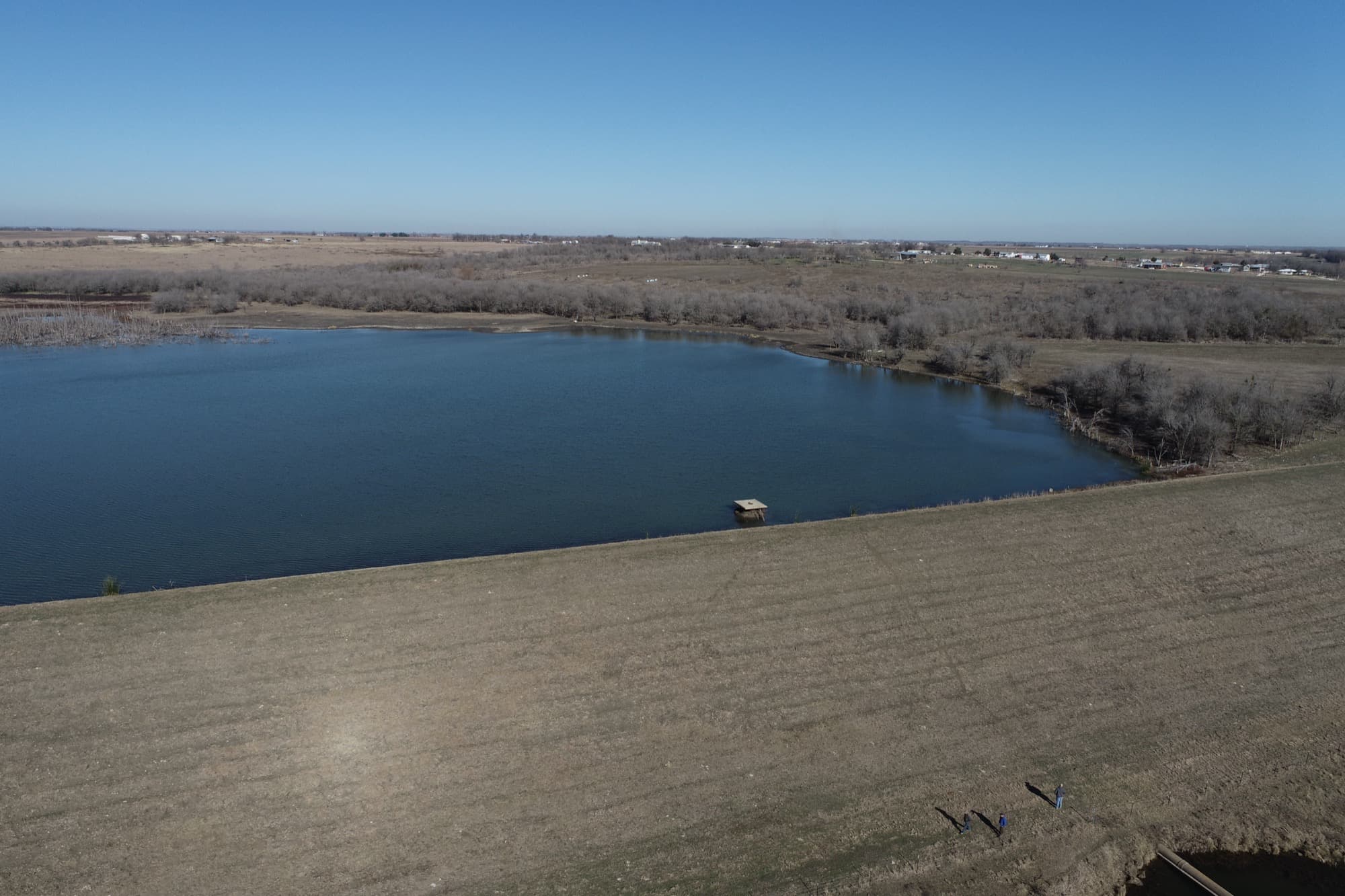 Aerial view of Lower Brushy Creek WCID Site 3 — a grass-covered earthen flood control embankment holding back its reservoir, with homes and open country beyond.