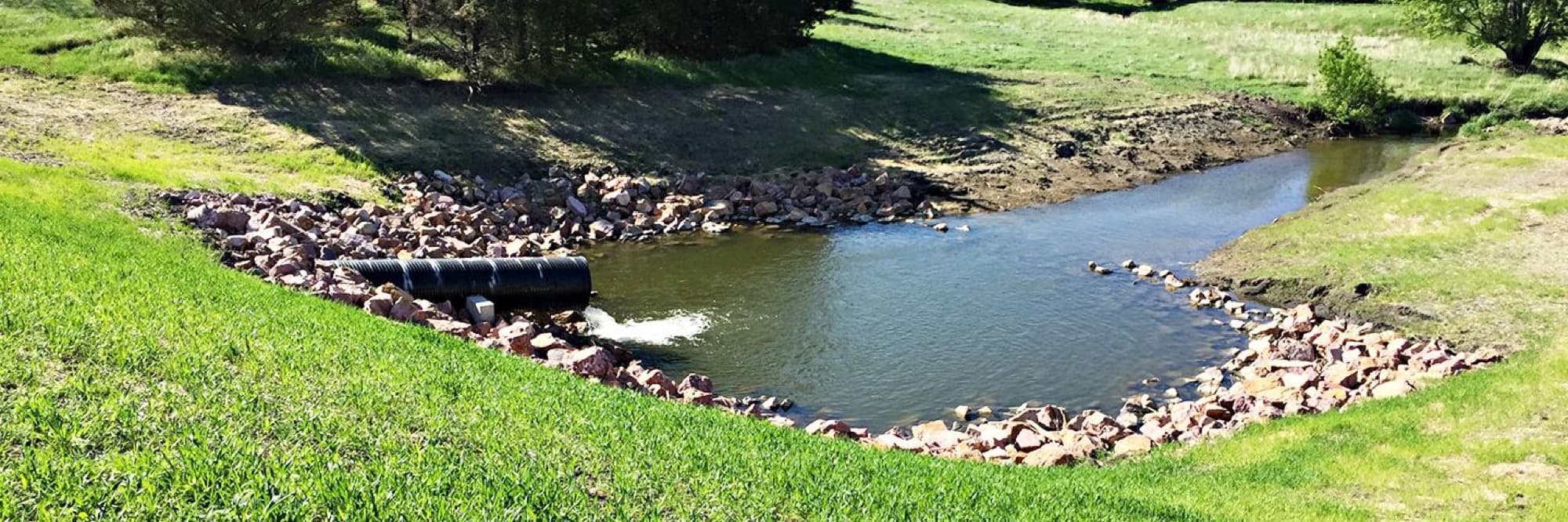 A principal spillway outlet pipe discharging into a rock-lined plunge pool below a grassed embankment.