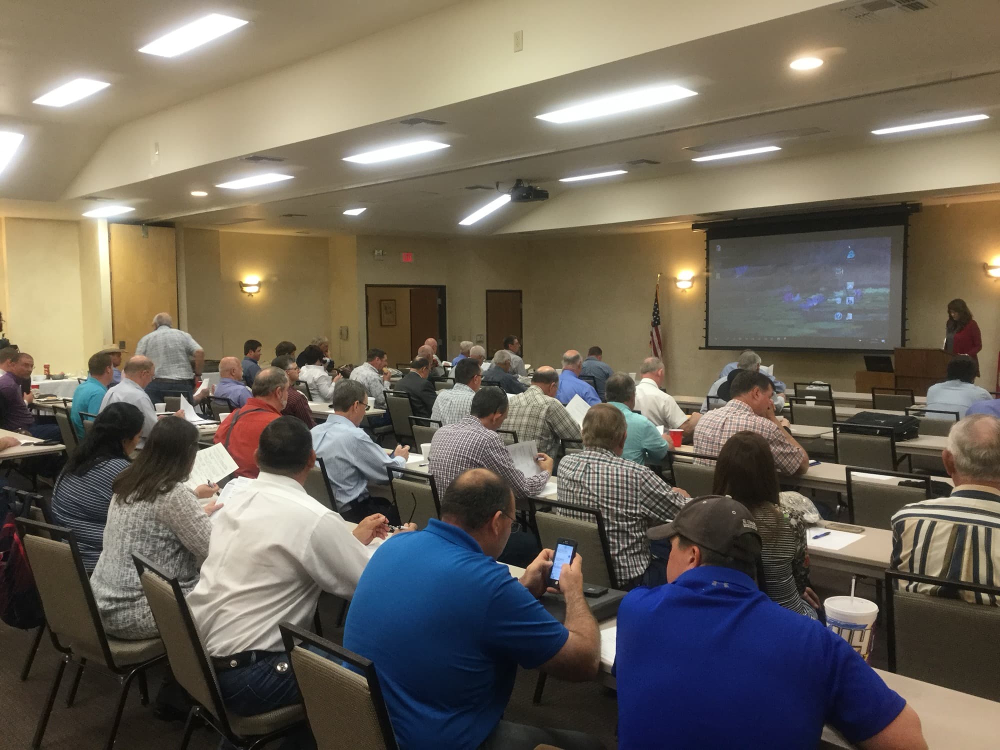 Attendees seated at tables during a TAWS annual meeting, with a presenter at the podium beside a projection screen.