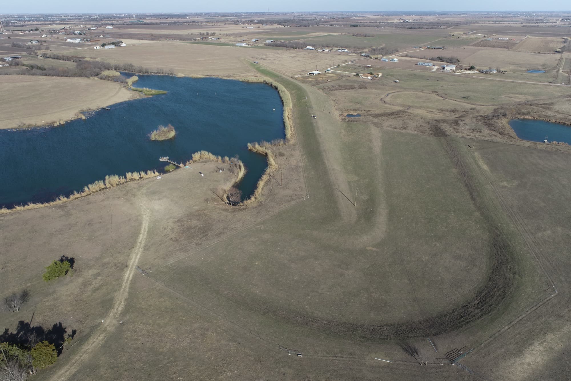 Aerial view of Upper Brushy Creek WCID Site 29, an earthen flood control embankment holding back its reservoir, with houses and farmland immediately downstream.